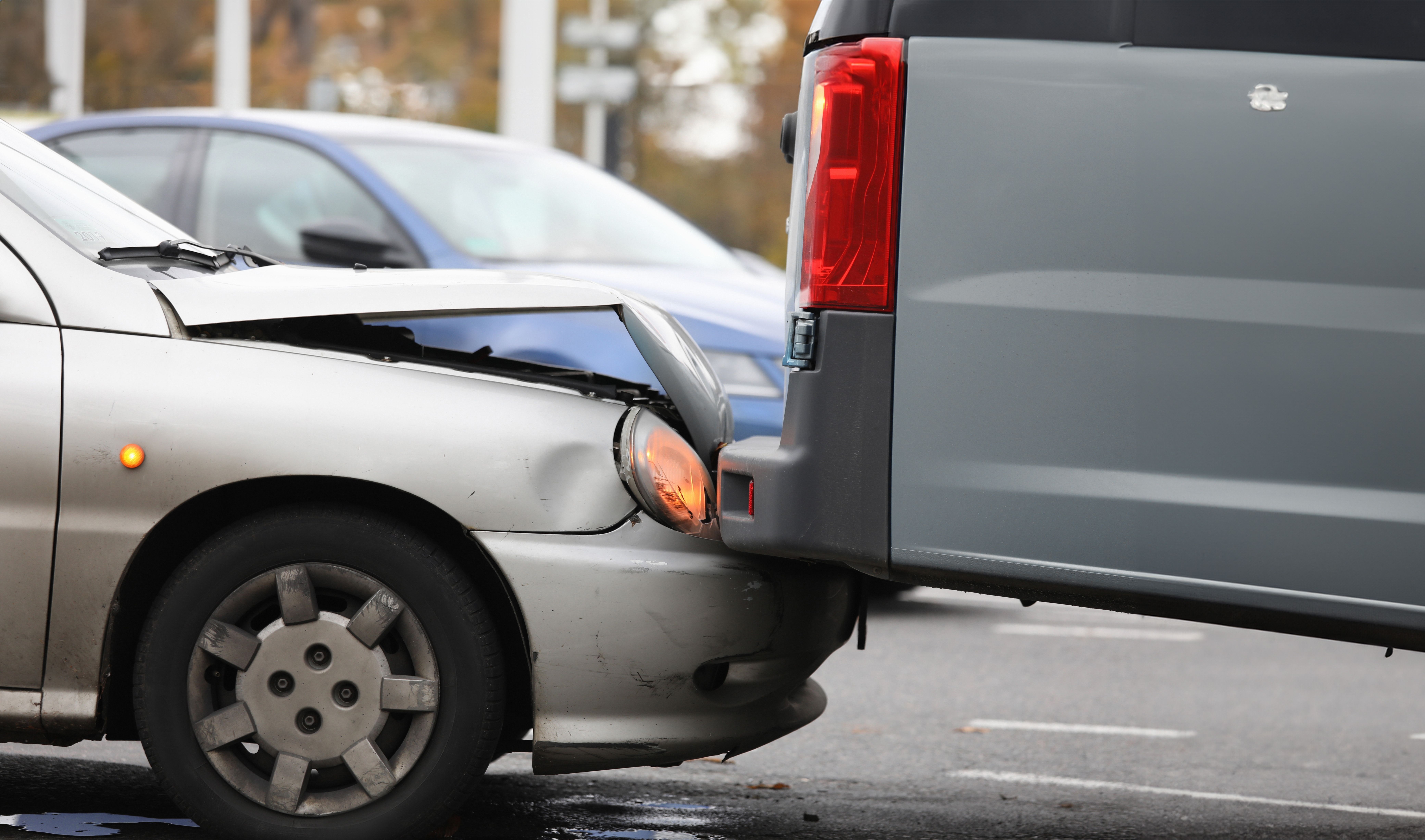 A silver car's damaged front end rests against the rear of a gray van after a collision.