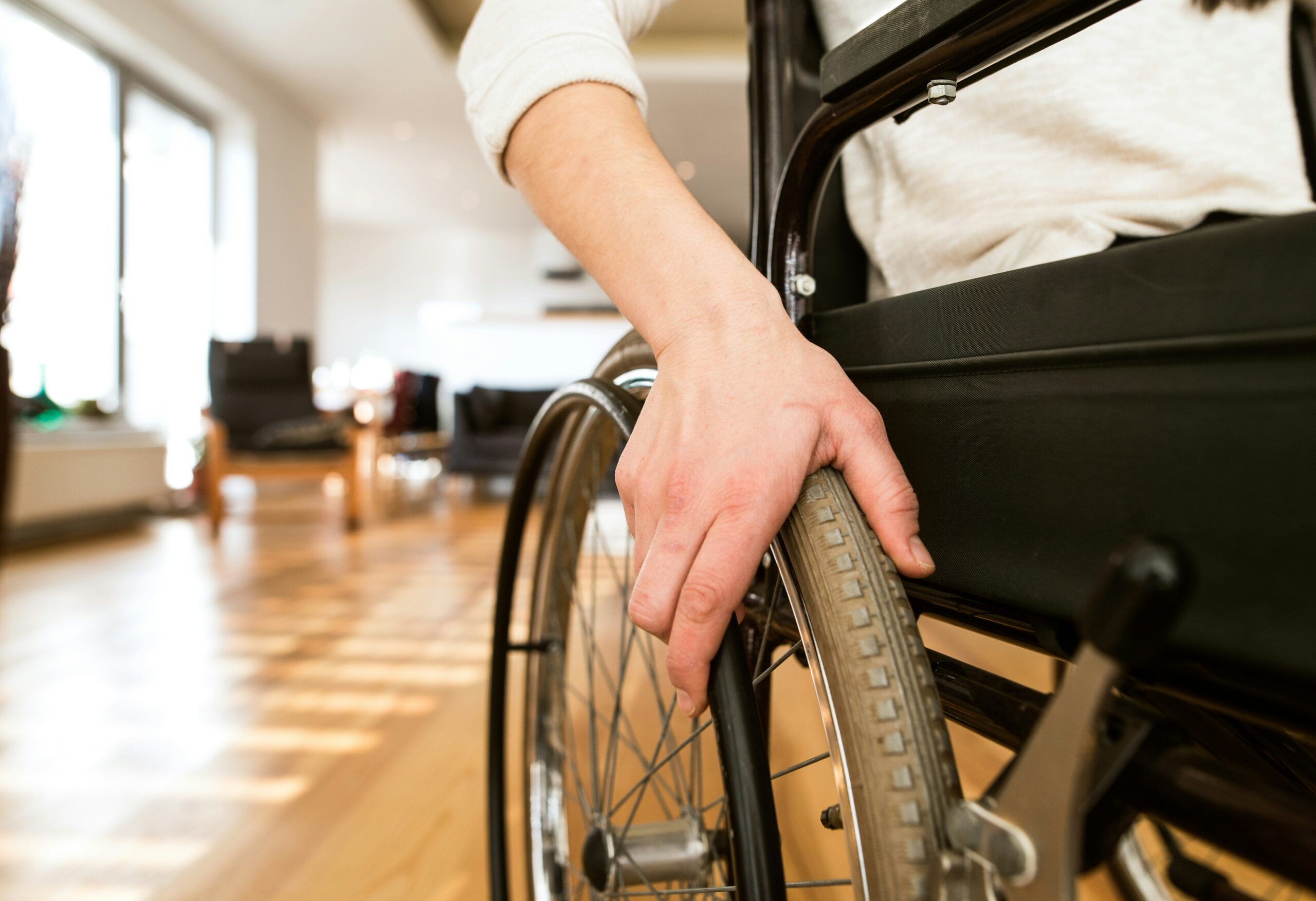 A close-up of a person’s hand gripping the wheel of a wheelchair indoors, with warm light reflecting off the wooden floor and a living room setting softly blurred in the background.