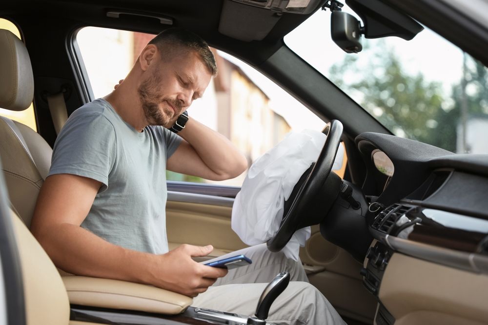 El conductor está sentado en el asiento del conductor del coche, con el airbag desplegado, mirando su móvil y sujetándose el cuello con gesto de dolor tras un accidente.