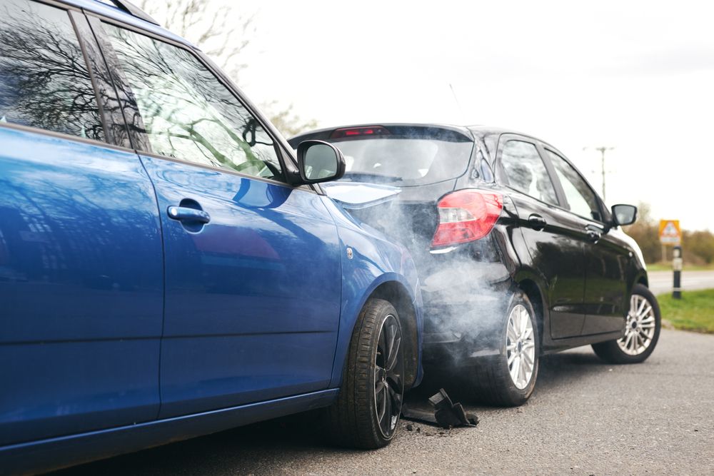 Blue car collided into the back of a black car with smoke rising after a rear-end accident on the road