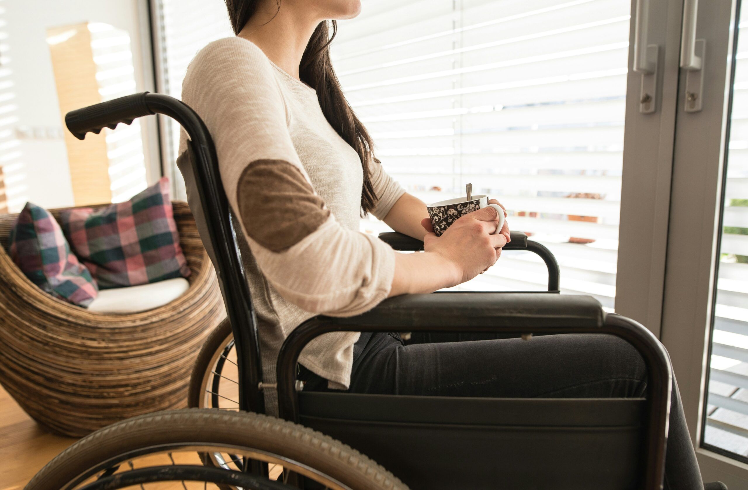 A young woman sitting in a wheelchair near large windows, holding a patterned mug with both hands and looking outside, with soft natural light coming through the blinds and a cozy chair with colorful cushions in the background.