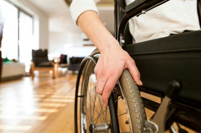 A close-up of a person’s hand gripping the wheel of a wheelchair indoors, with warm light reflecting off the wooden floor and a living room setting softly blurred in the background.