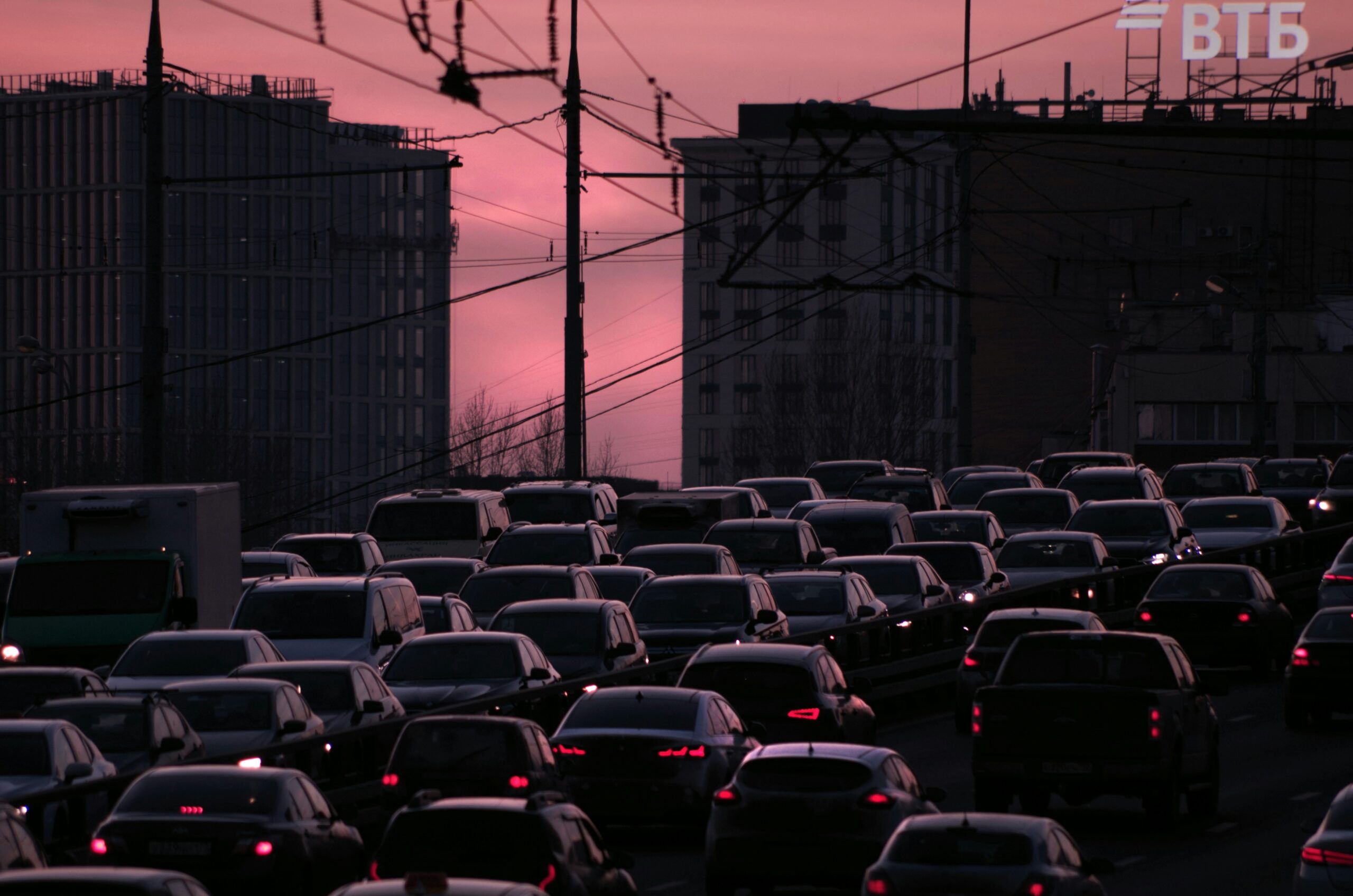 Heavy urban traffic congestion at dusk, with rows of cars packed closely together beneath overhead power lines, silhouetted buildings on both sides, and a pink-purple sunset sky casting a dim glow over the city scene.
