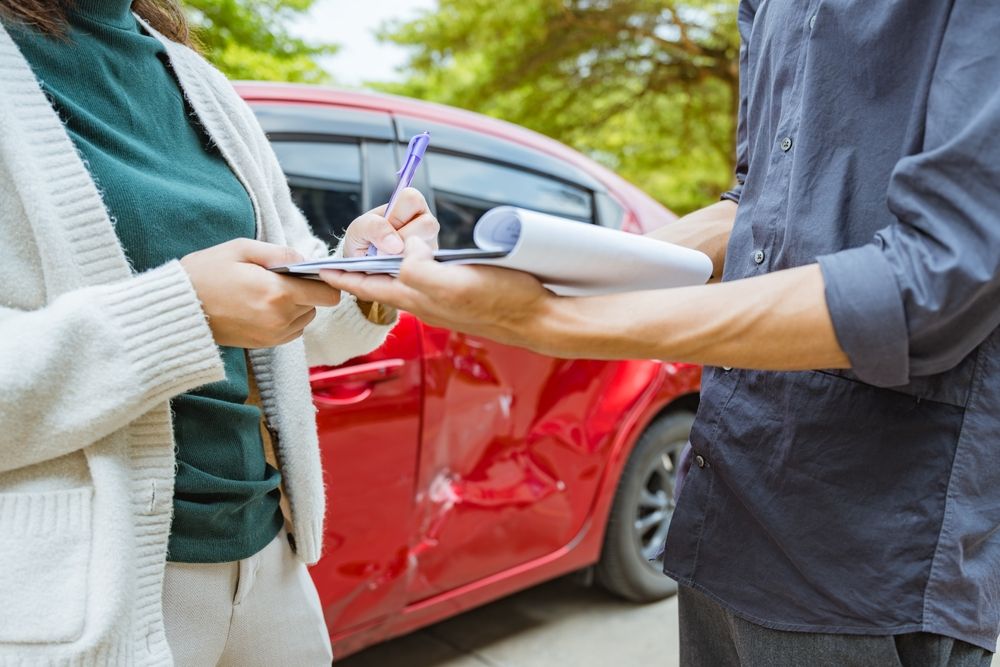 Two people exchanging information on a clipboard beside a damaged red car after an accident