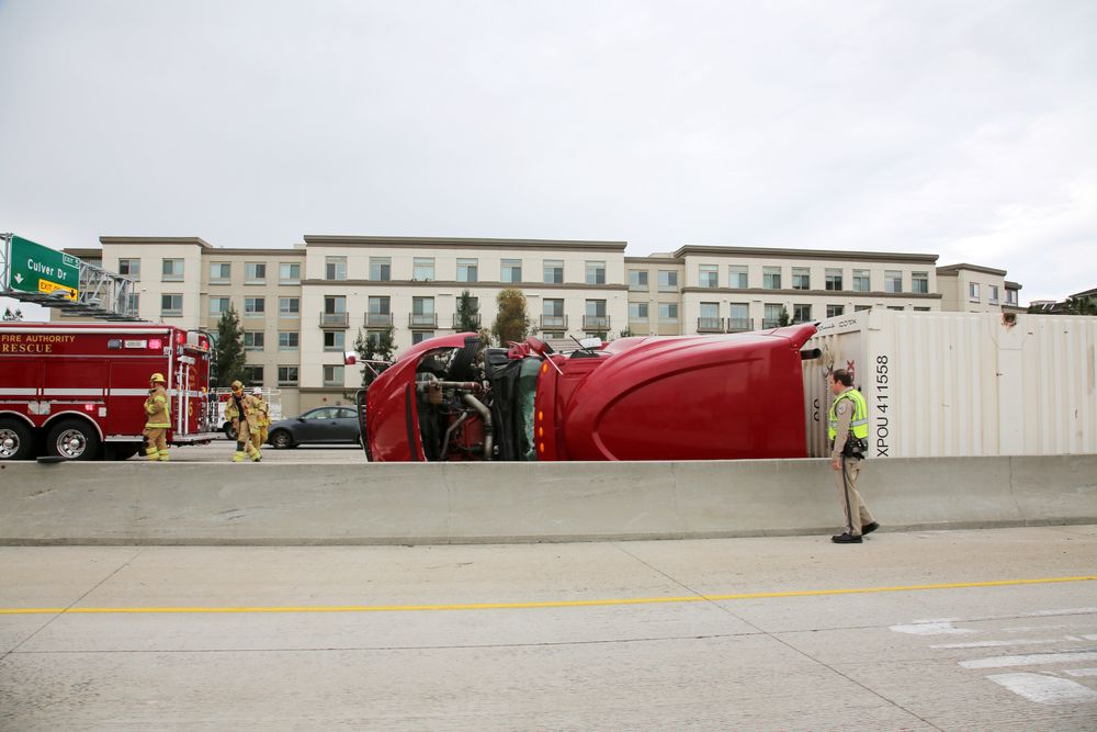 Overturned red semi-truck on a highway with firefighters and a police officer responding at the scene