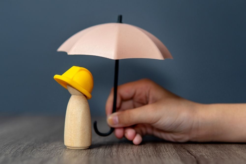 Hand holding a small umbrella over a wooden figure wearing a yellow hard hat