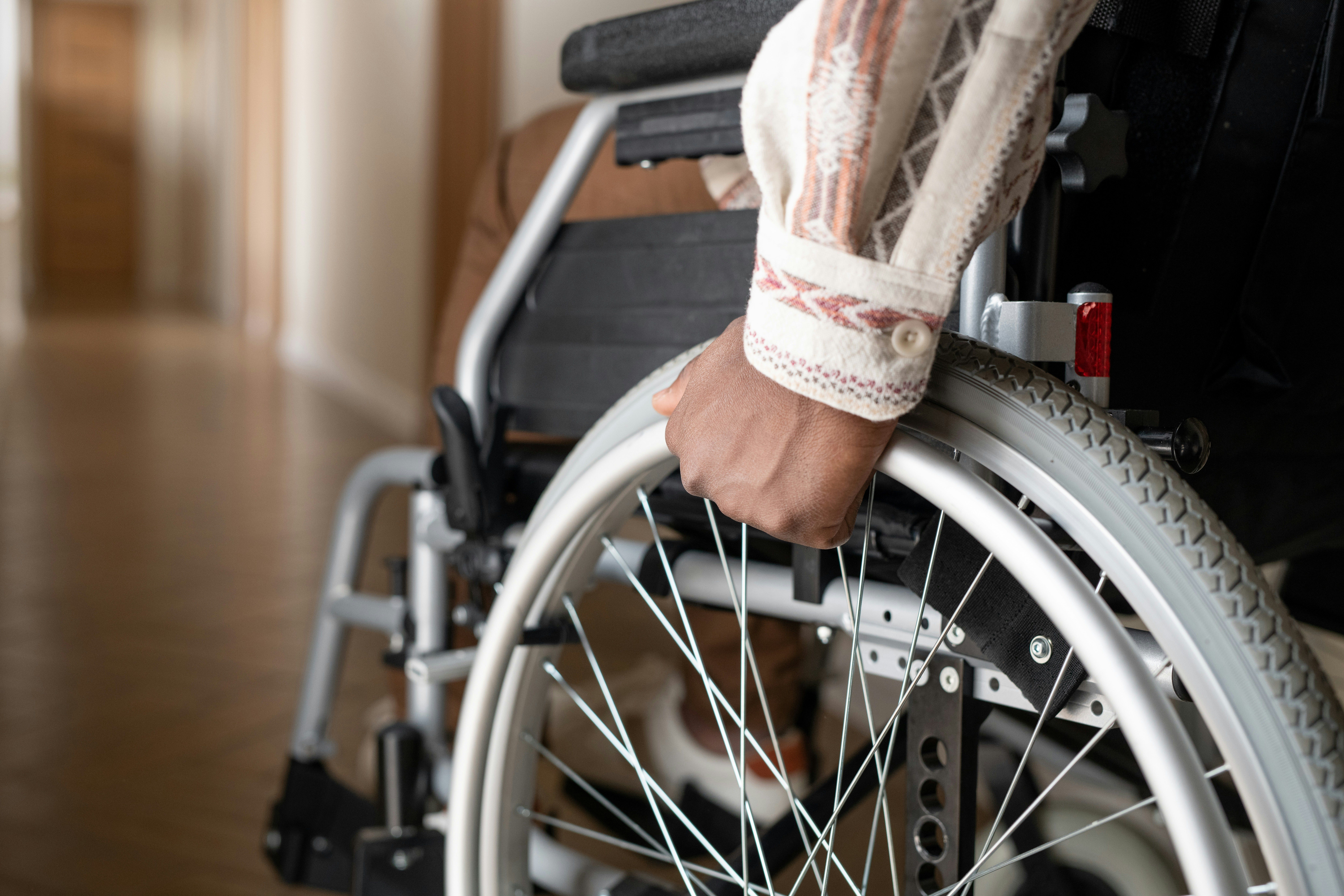 Close-up of a person’s hand gripping the wheel of a manual wheelchair in an indoor hallway, showing detailed spokes, tire tread, and the wheelchair frame, with the person seated and wearing patterned clothing, emphasizing mobility and physical injury recovery.