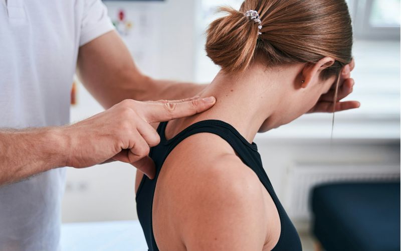 A clinician gently presses along the back of a woman’s neck as she leans forward, assessing muscle tension or pain during a physical examination in a bright medical setting.