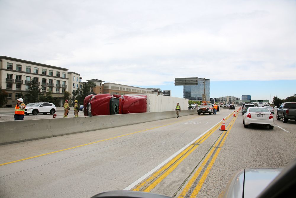 Un camión articulado rojo volcado y tumbado de costado bloquea la mediana de una autopista, mientras los servicios de emergencia acuden al lugar y el tráfico sigue circulando.