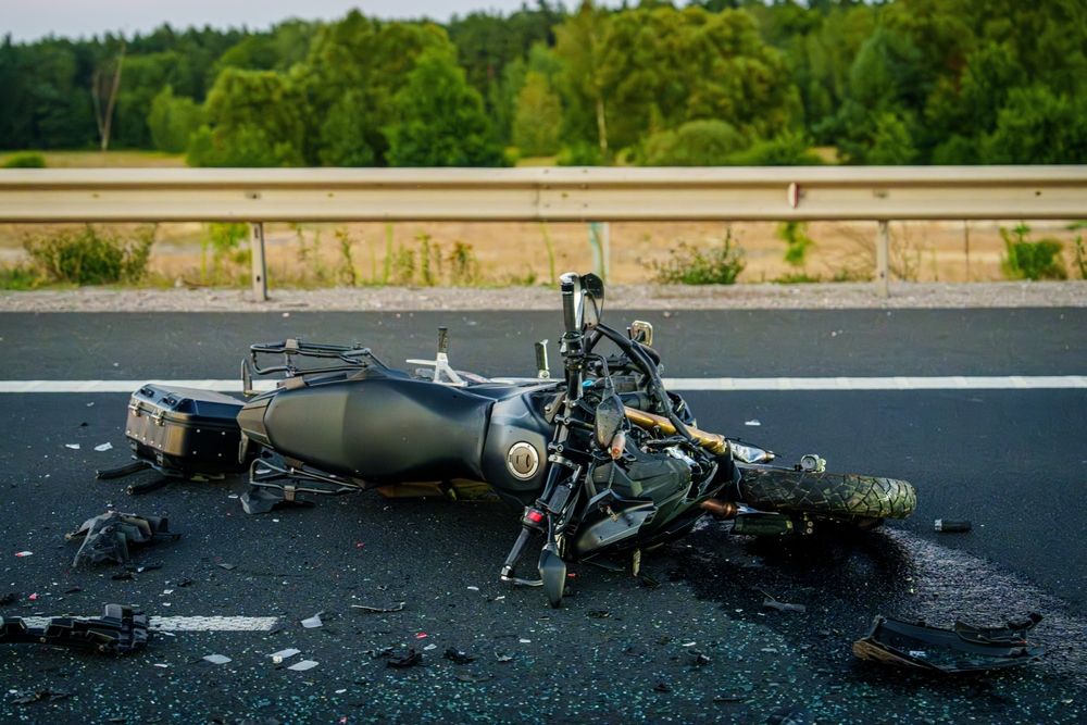 Motorcycle lying on its side after a crash on a highway, debris scattered around with a guardrail in the background.