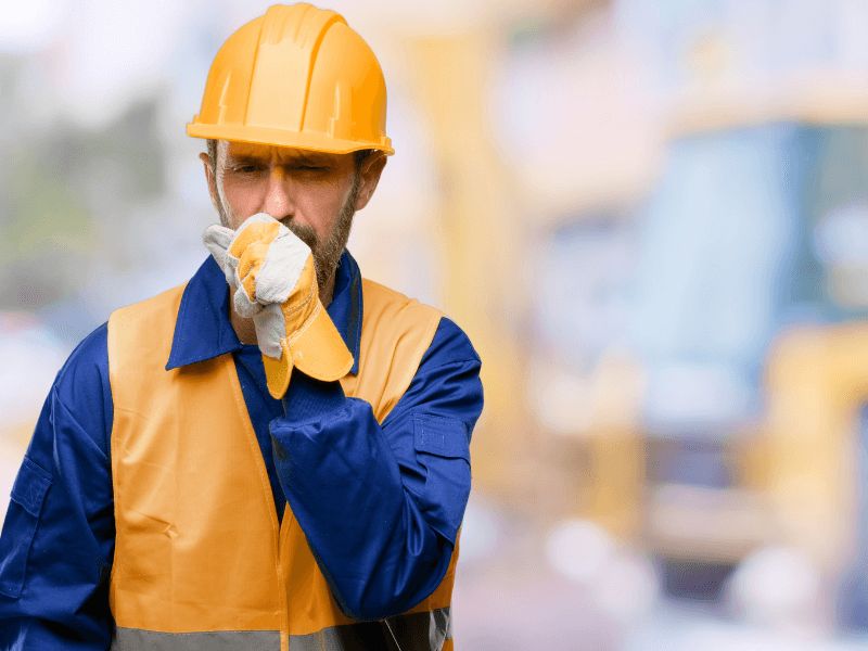 Man wearing hard hat coughing into gloved hand