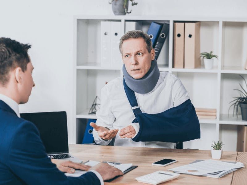 man in neck brace and arm sling talking to man in suit at desk