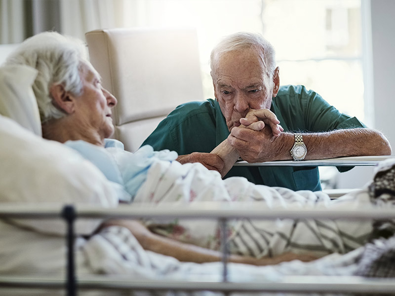 elderly man kissing his wifes hand while she is in a hospital bed