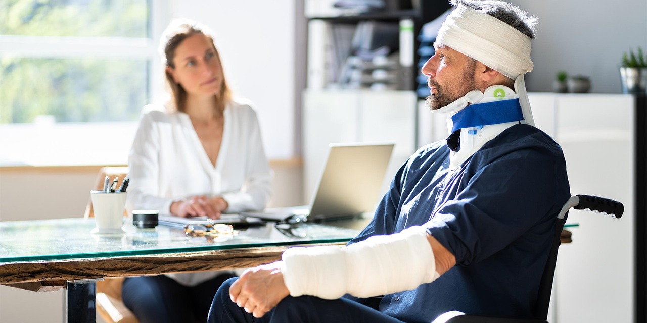 A man in a wheelchair with a cast on his arm, looking determined and focused, is across a glass desk from a woman who looks at him with compassion
