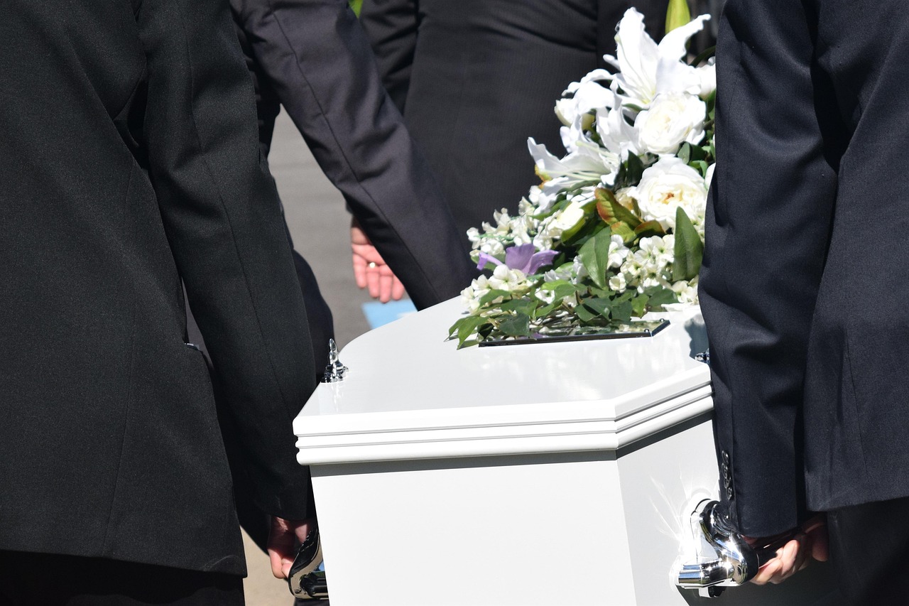 A solemn funeral procession featuring a casket being carried by mourners along a tree-lined path.