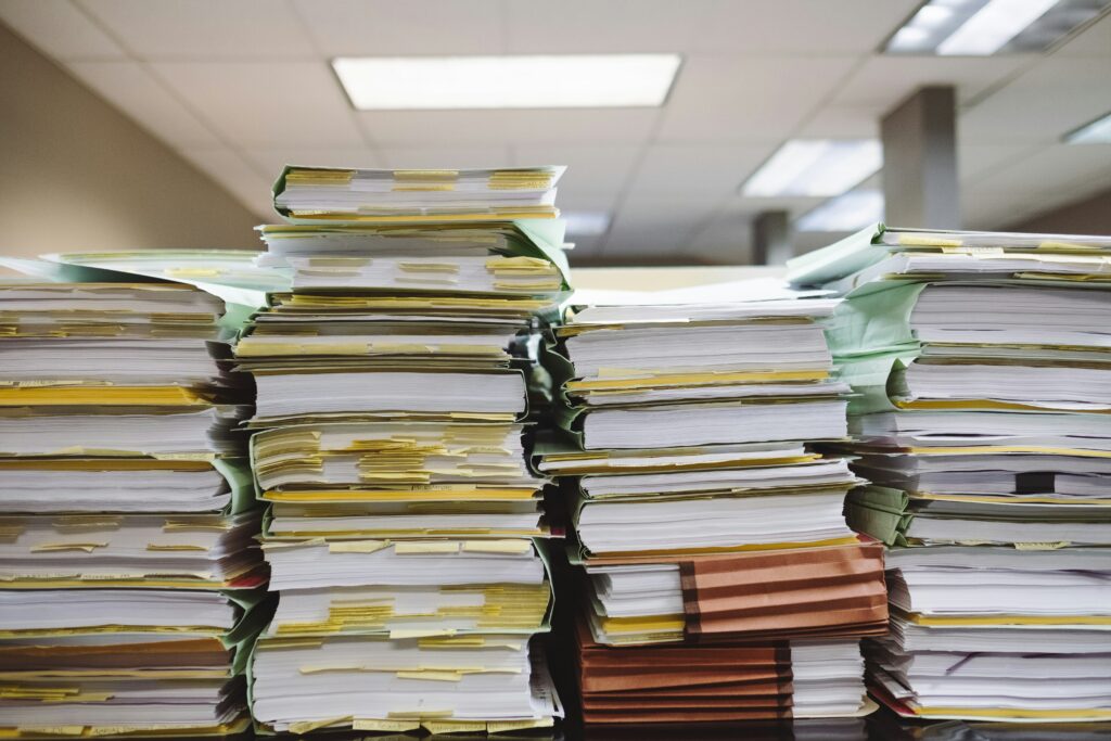 Una pila de papeles y carpetas repletas de documentos descansa sobre un escritorio en un edificio de estilo administrativo con duras luces que se oyen por encima.