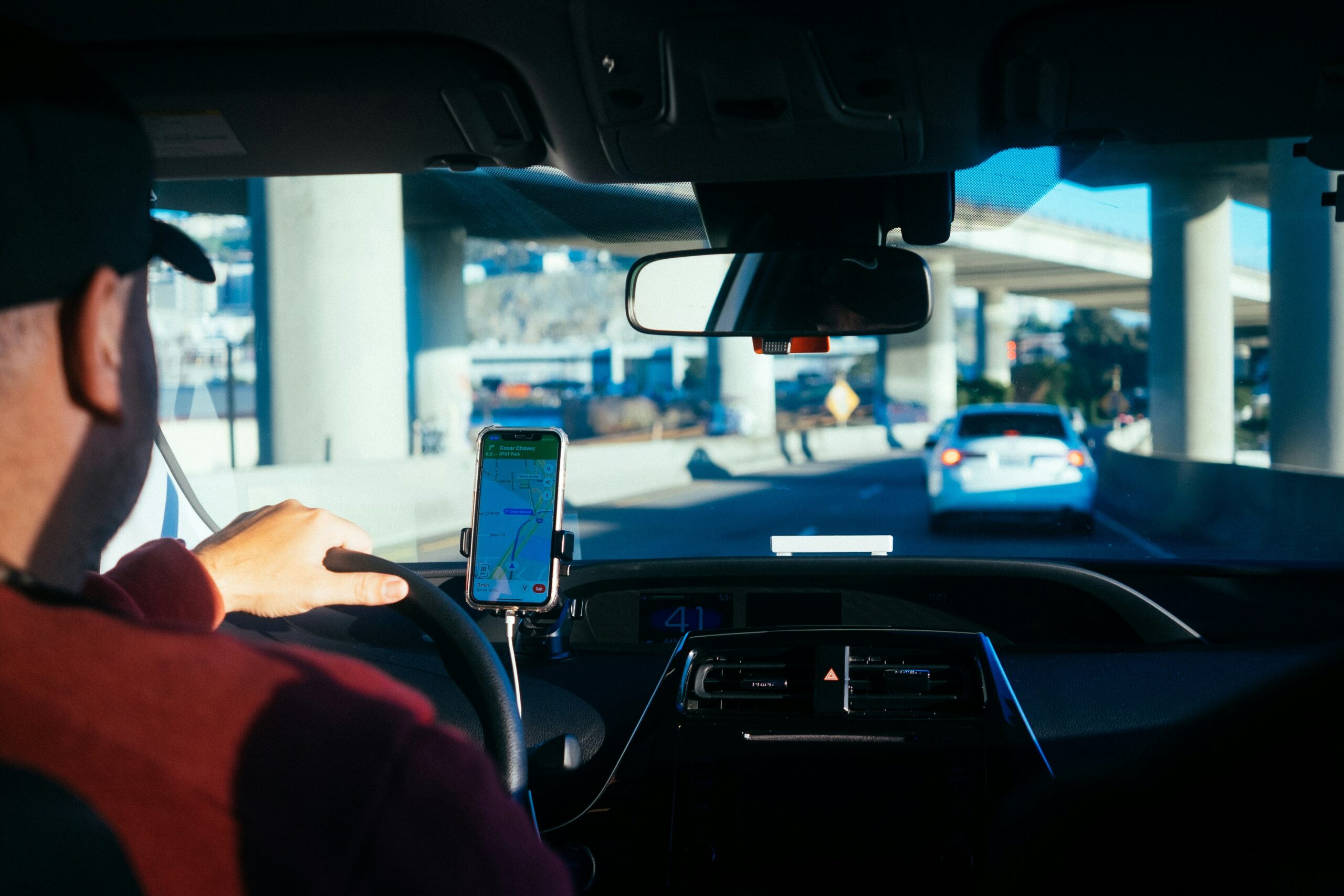 a rideshare driver in a red shirt and a black cap looks out at the cars ahead of him, with his phone's GPS in view