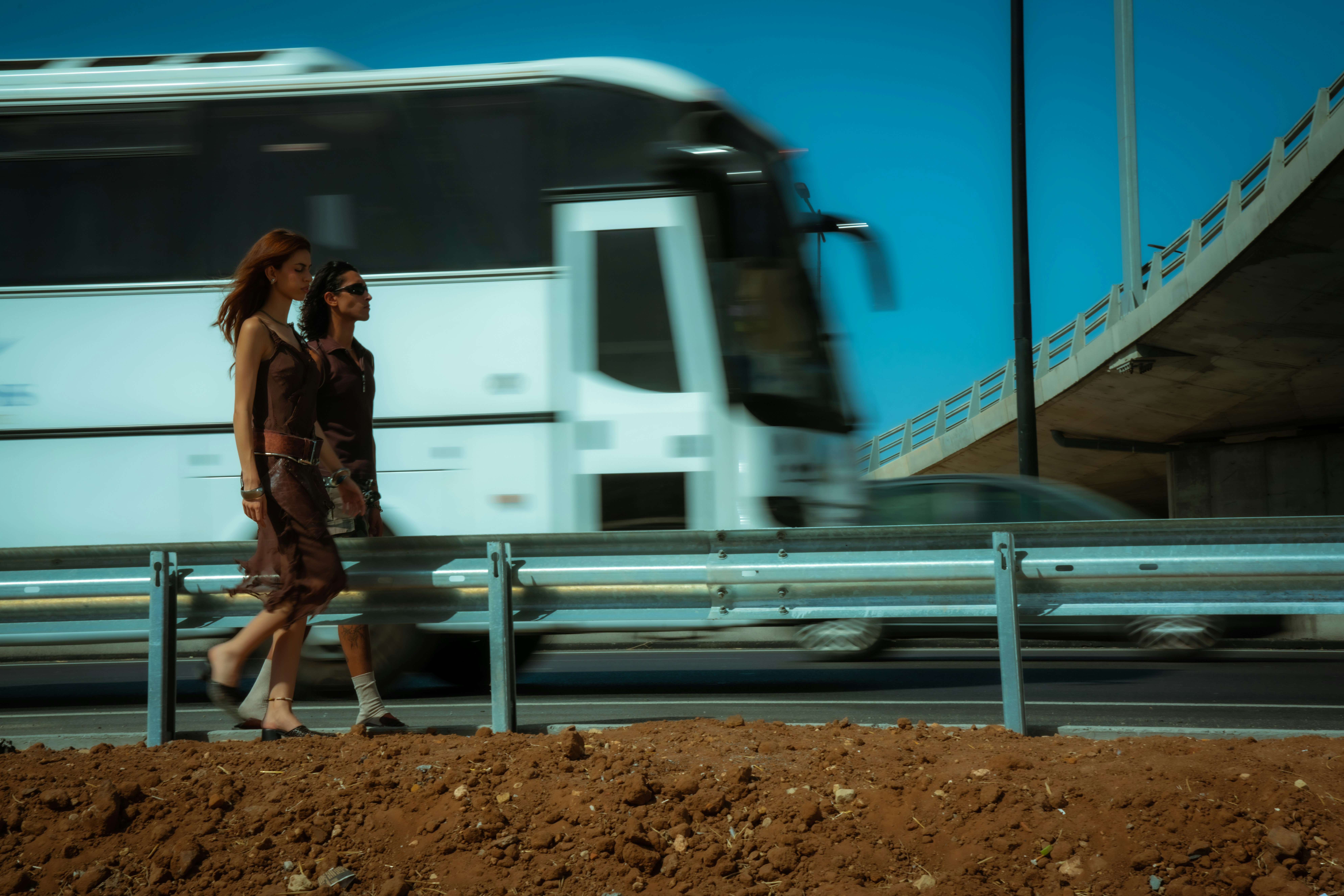two people walking on a highway while a bus drives past