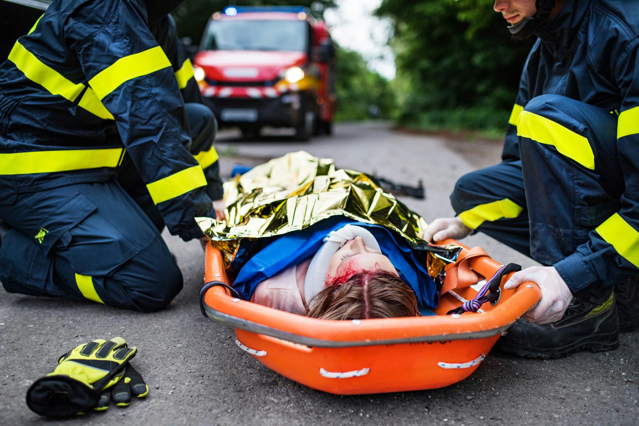 Two emergency responders kneeling on a road secure an injured person onto an orange stretcher, with the patient covered in a thermal blanket and showing visible facial injuries while an ambulance is parked in the background.