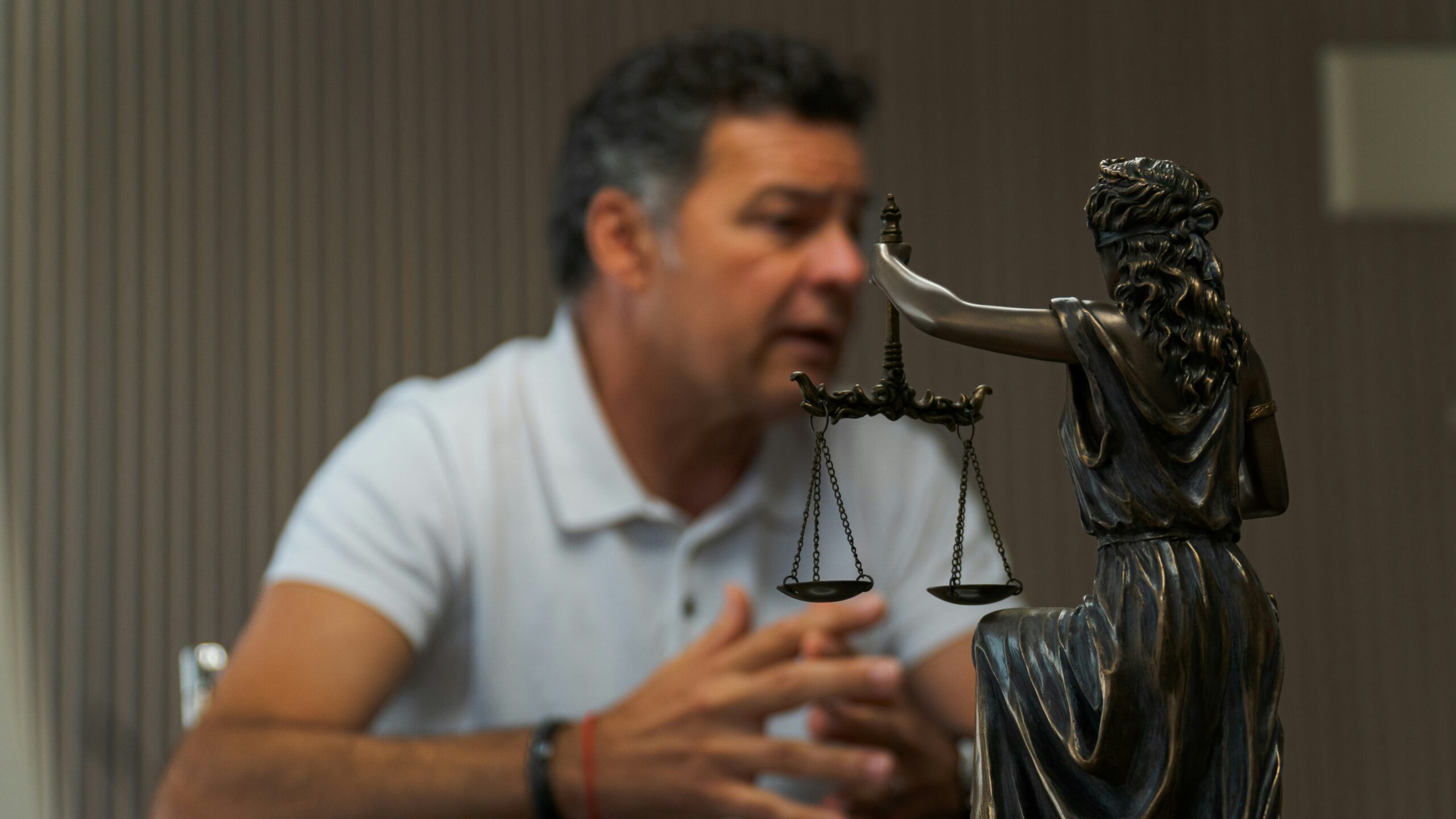 a man sits at a table in the background, clearly speaking to someone we cannot see, with a lady justice statue in the foreground