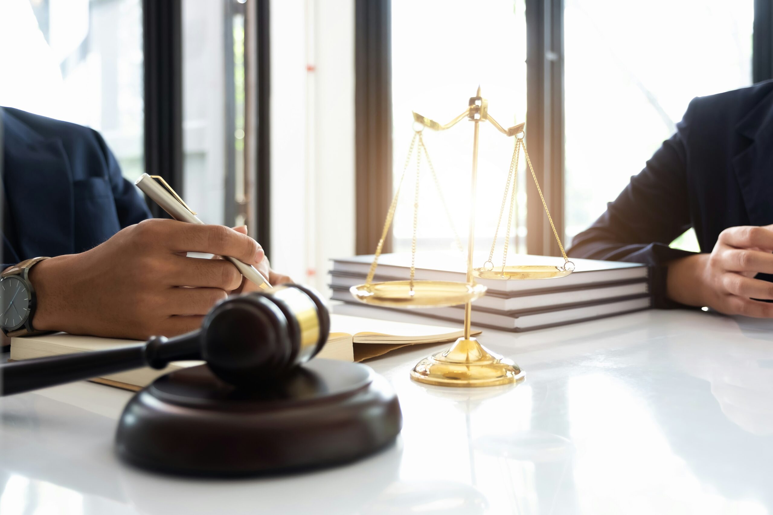 A close-up view of two professionals in suits sitting at a desk, with one writing in a notebook while legal symbols such as a judge’s gavel and golden scales of justice sit prominently in the foreground beside a stack of books.
