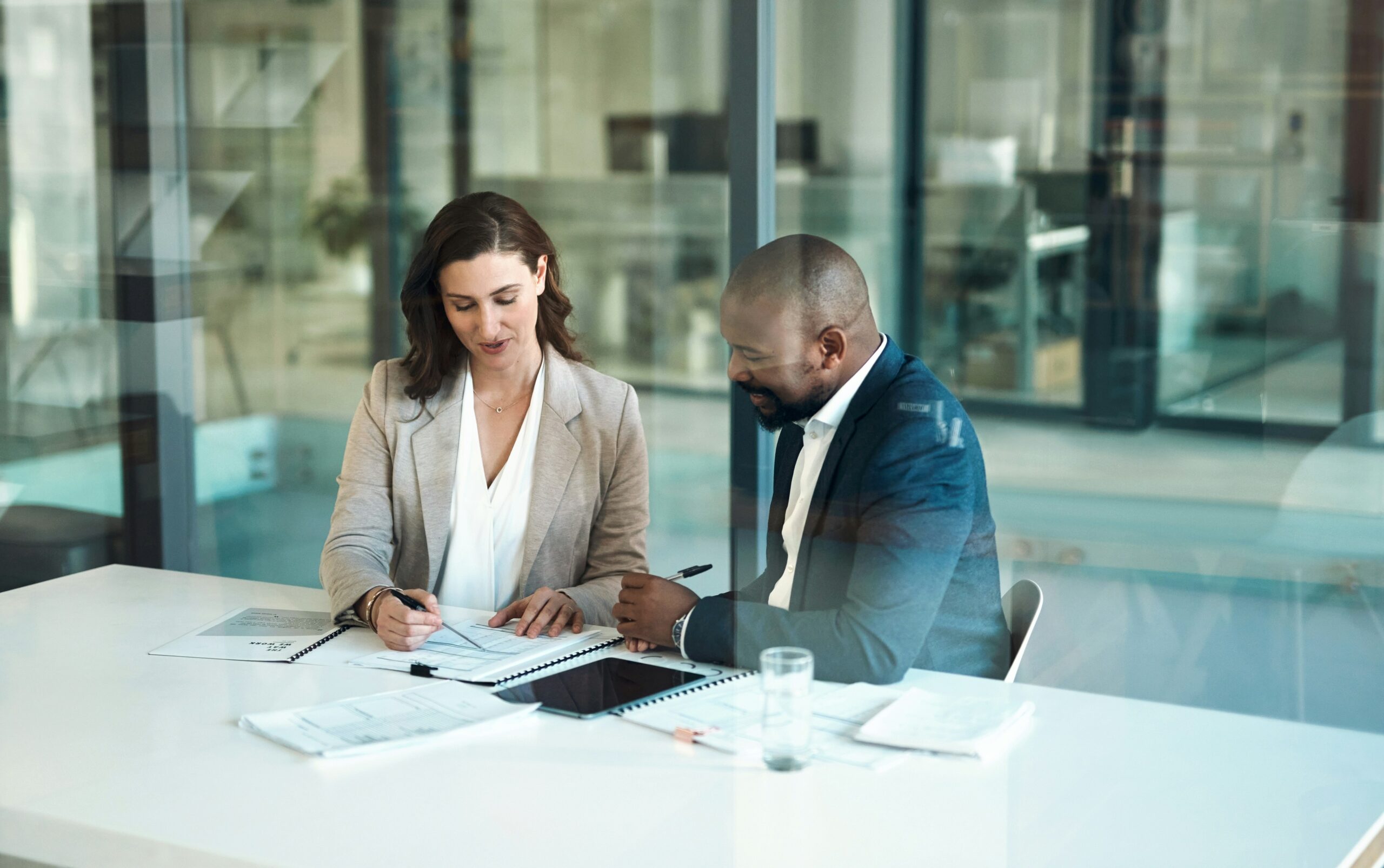 Two professionals sitting together in a modern glass-walled office, reviewing documents at a white table, with one person pointing to a page while the other makes notes.