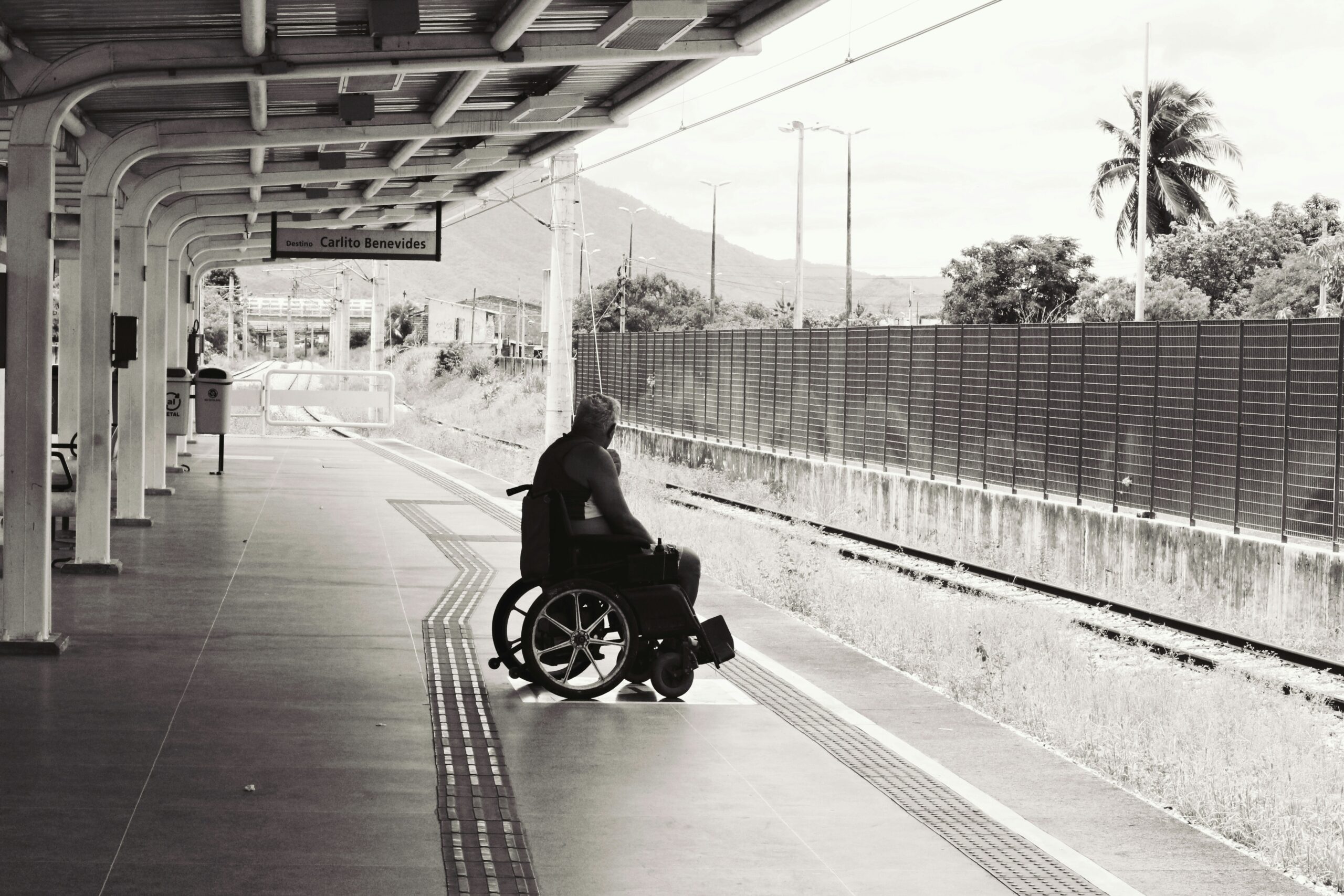 A man in a wheelchair waiting alone on a quiet train platform, positioned near the edge of the tracks under the covered station roof, with distant mountains and palm trees visible in the background.