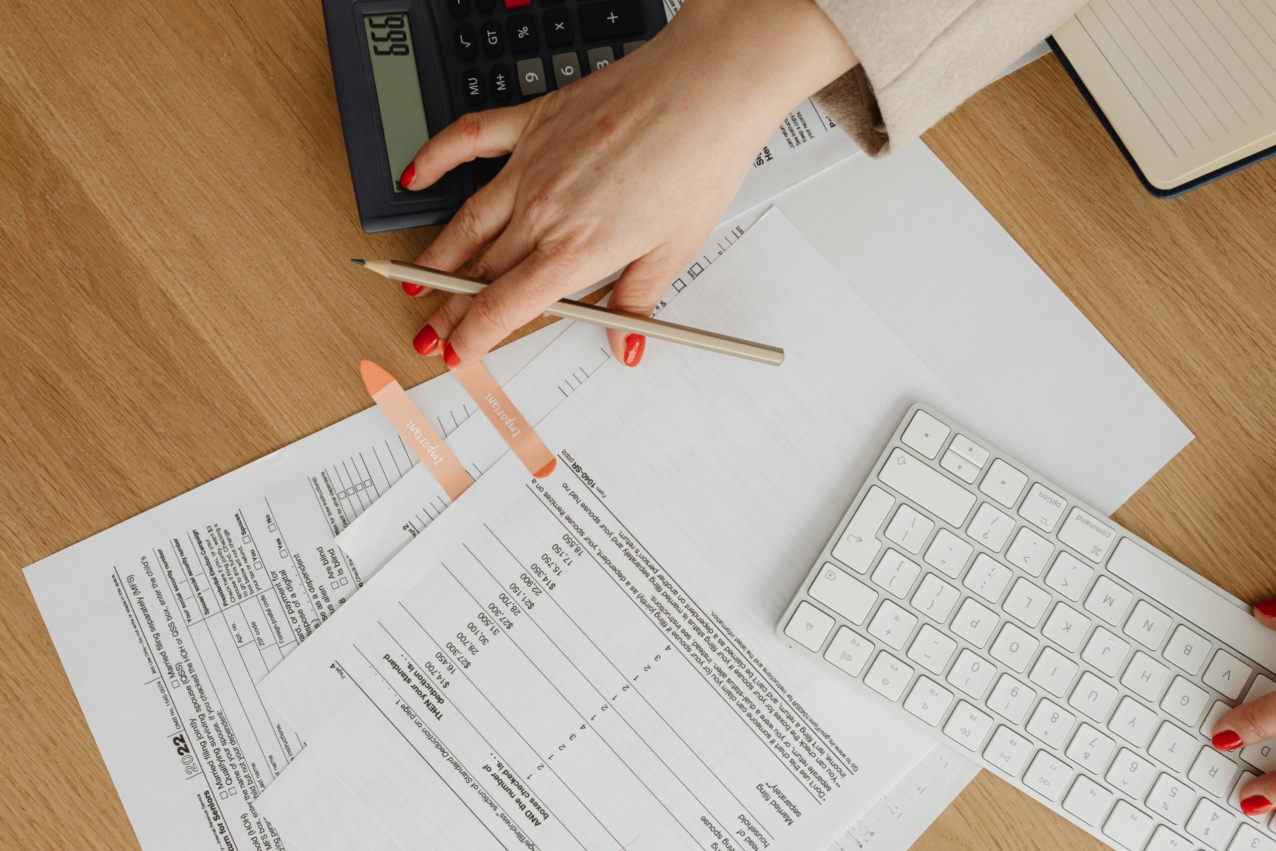 A close-up of a person reviewing financial documents at a desk, with tax forms spread out, a calculator nearby, and one hand holding a pencil while the other rests on a white keyboard.