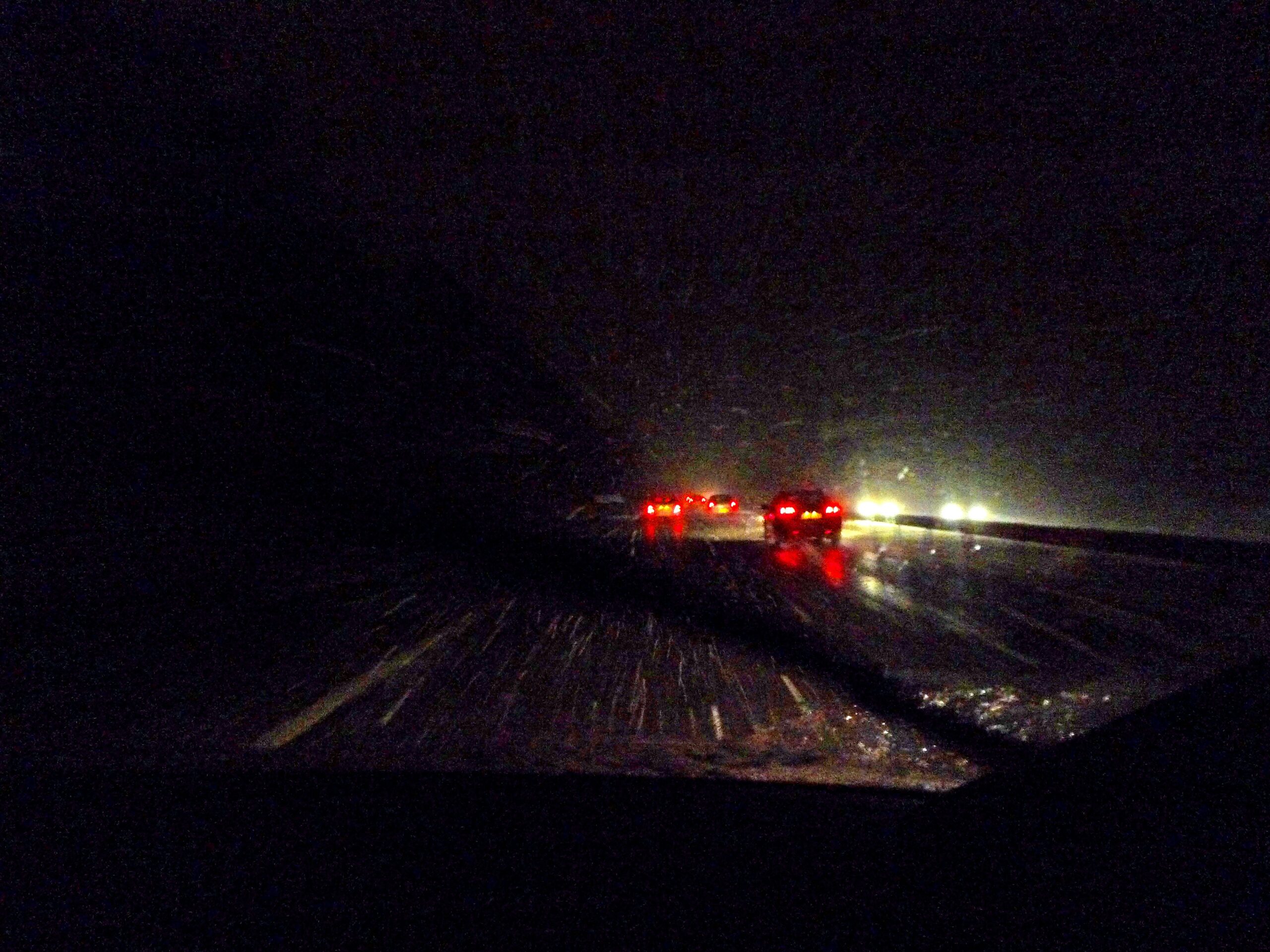 Nighttime highway scene viewed through a windshield during snowfall, with snow streaking across the road, dark surroundings, wet reflective pavement, and several vehicles ahead marked by glowing red taillights and distant oncoming headlights.