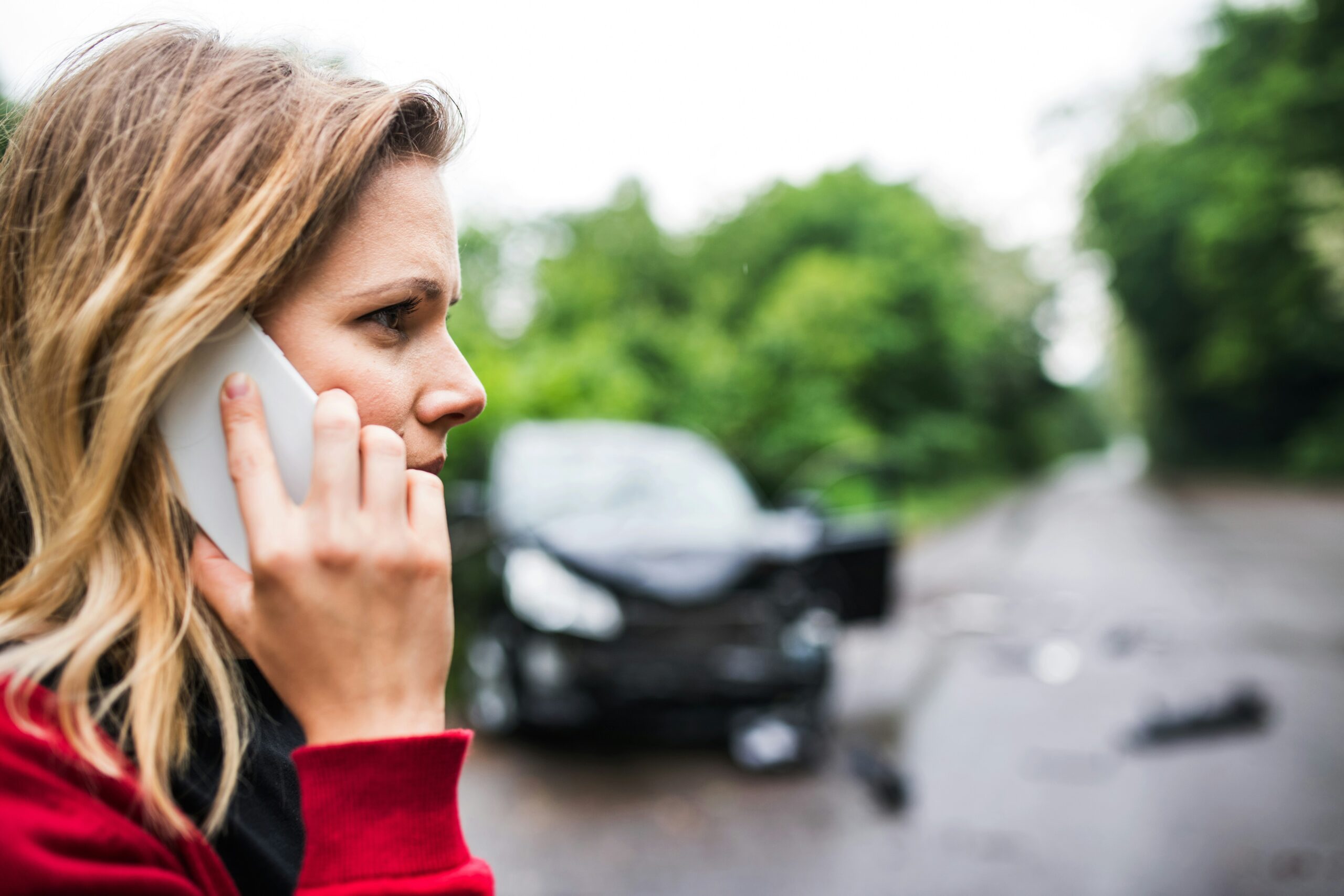 Side profile of a woman holding a smartphone to her ear on a tree-lined road, appearing tense and focused, with a blurred damaged car and scattered debris visible in the background, suggesting a recent traffic accident scene.
