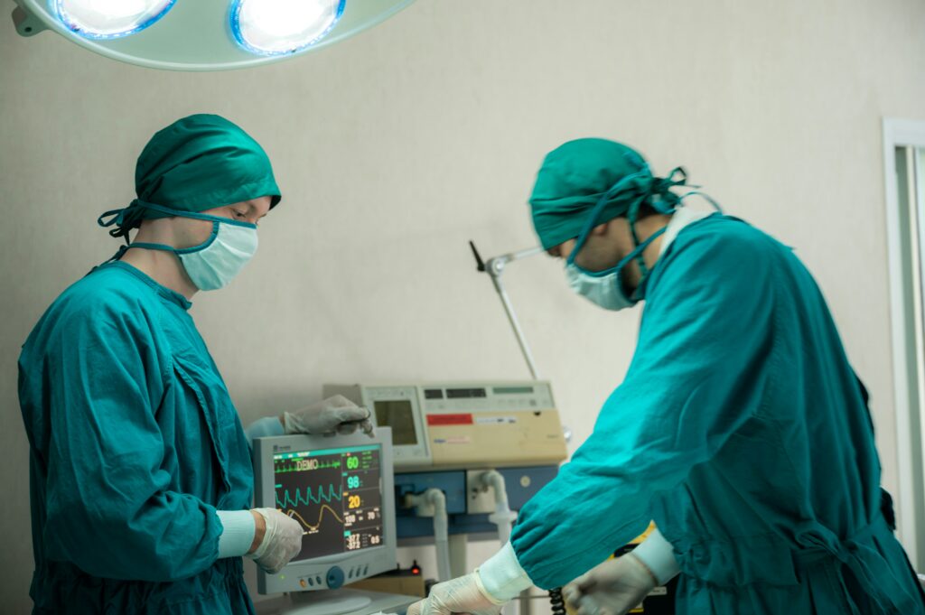 Dos cirujanos vestidos con batas quirúrgicas verdes, gorros, mascarillas y guantes se encuentran en un quirófano bajo luces quirúrgicas, concentrados en un paciente que no aparece en el encuadre mientras controlan los signos vitales que se muestran en un monitor médico situado junto a ellos.