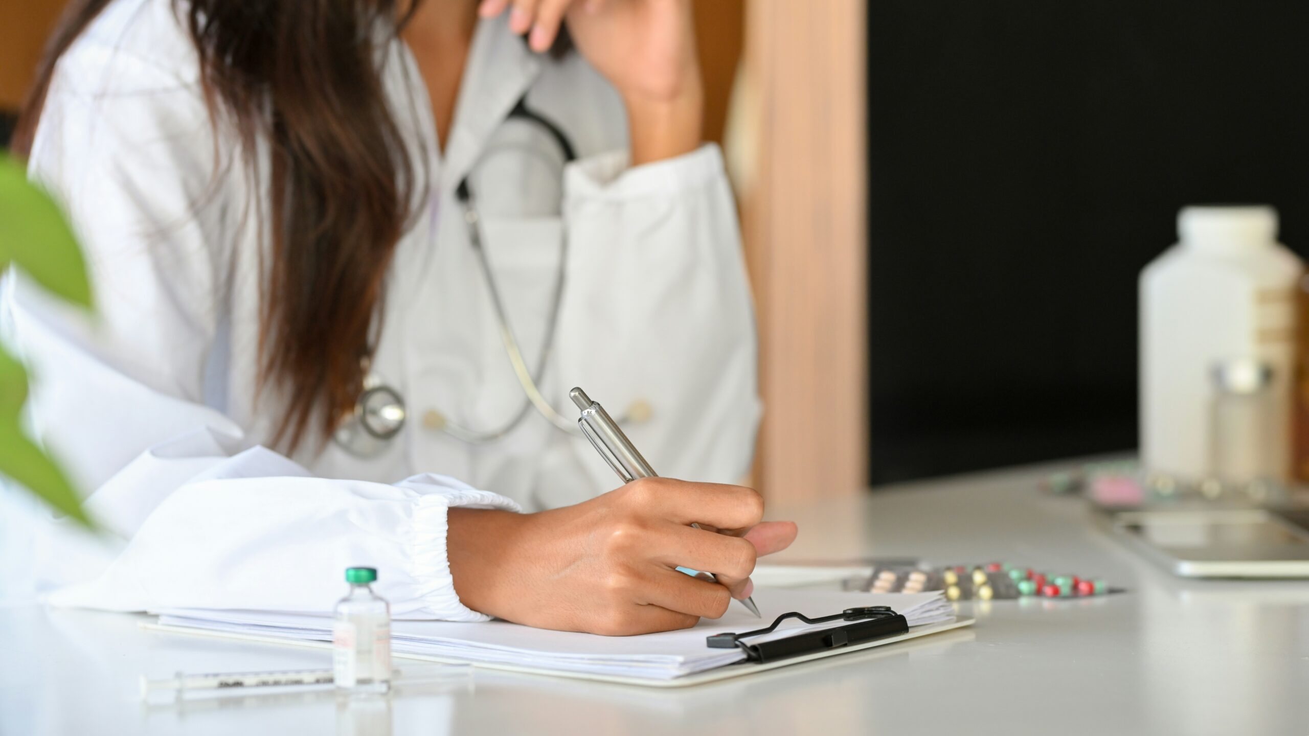 A medical professional in a white lab coat with a stethoscope writes on a clipboard at a desk, with prescription pill bottles, a blister pack of medication, and a small vial visible nearby in a clinical office setting.