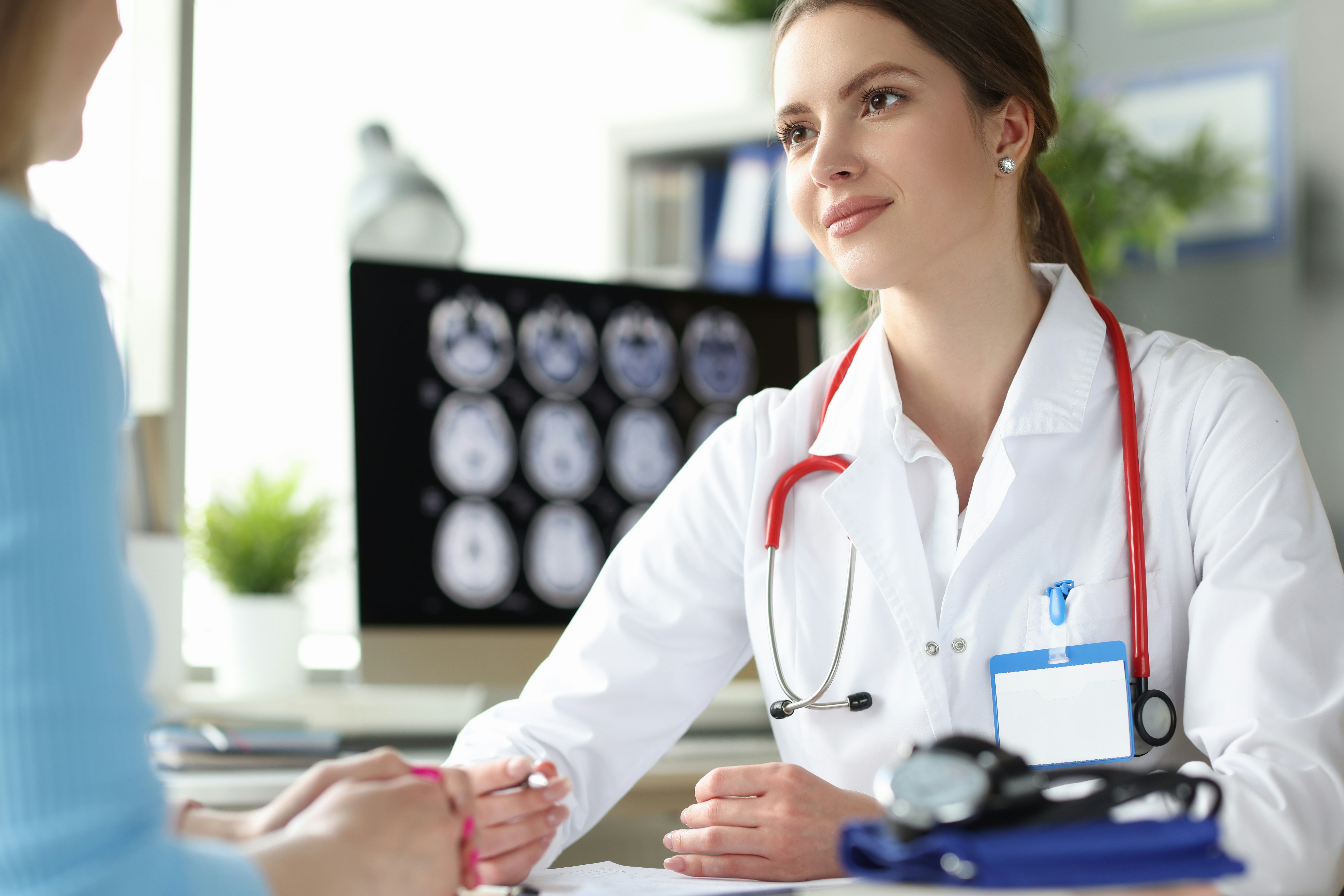 A doctor in a white coat listens attentively to a patient during a consultation, with a stethoscope around her neck and diagnostic brain scans visible on a monitor in the background.