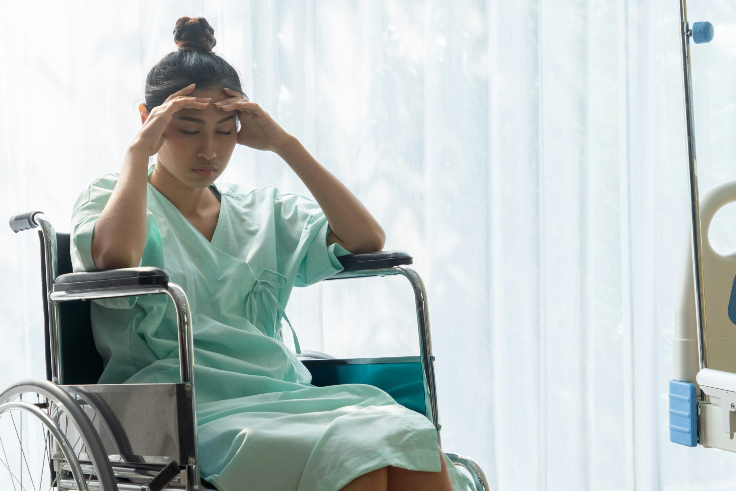 A woman wearing a hospital gown sits in a wheelchair beside a hospital bed, holding her head with both hands and looking down with eyes closed, suggesting pain, stress, or exhaustion, in a bright hospital room with sheer curtains and medical equipment visible nearby.