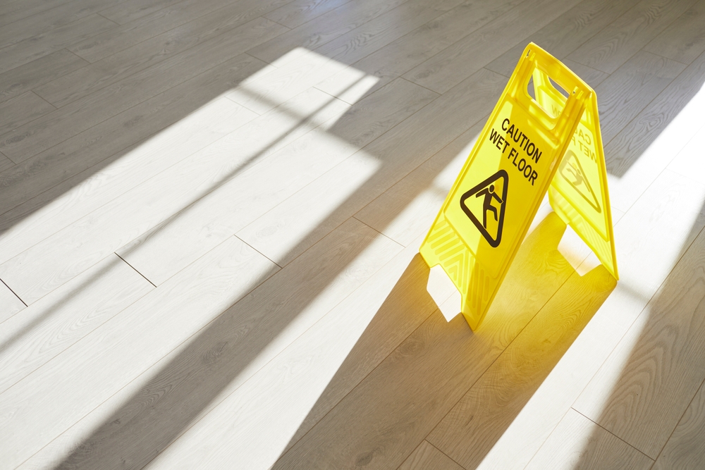 Yellow "Caution: Wet Floor" sign standing on a hardwood floor with sunlight streaming across it.